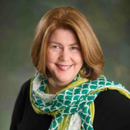 Theresa Schwerin posed headshot. She wears black with a green and yellow scarf around her neck. 