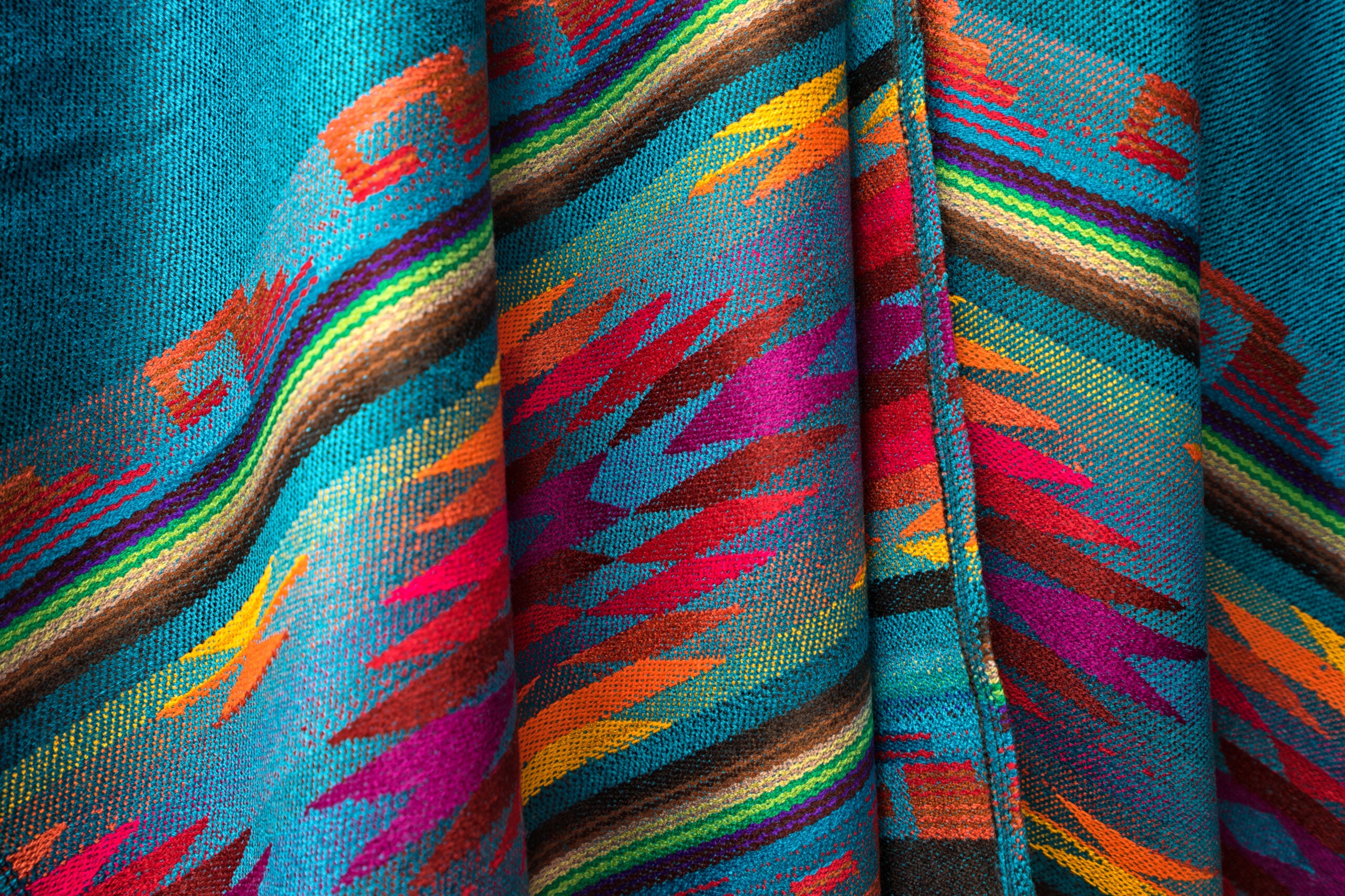 
		A close-up image of a vibrant, woven textile featuring a colorful and intricate pattern. The fabric includes various shades of turquoise, red, pink, orange, yellow, green, and brown, arranged in geometric and zigzag designs. The texture of the weave is visible, showcasing the detailed craftsmanship and the interplay of colors, creating a visually striking and dynamic appearance.		