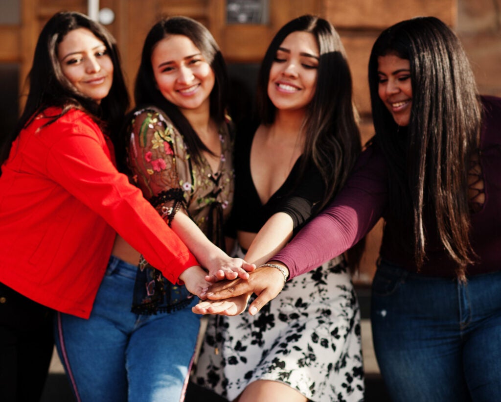 A group of four young women standing close together with their hands stacked in the center, symbolizing unity and friendship. They are smiling and appear to be enjoying each other's company. The women are dressed in casual, colorful clothing, with one wearing a red jacket, another in a floral blouse, the third in a black top and patterned skirt, and the fourth in a dark top. The background features a wooden door, suggesting an outdoor setting.